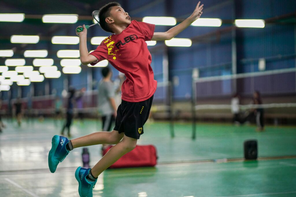 A young boy enthusiastically jumps to hit a badminton shuttlecock inside a sports hall.